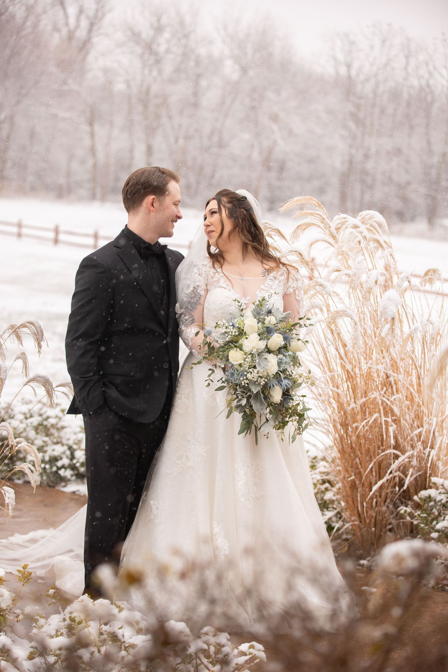 A romantic winter wedding scene with the couple standing near ornamental grasses in falling snow.
