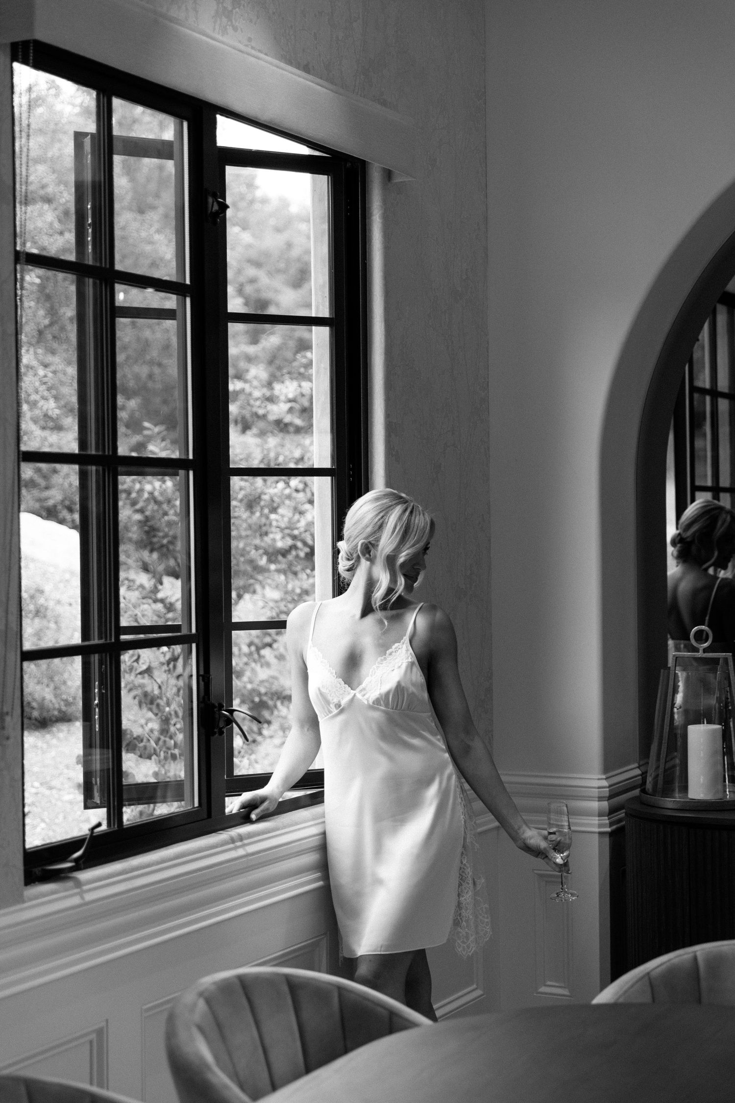 Black and white photograph shows a figure in a white dress looking out through a large window with grid panes.