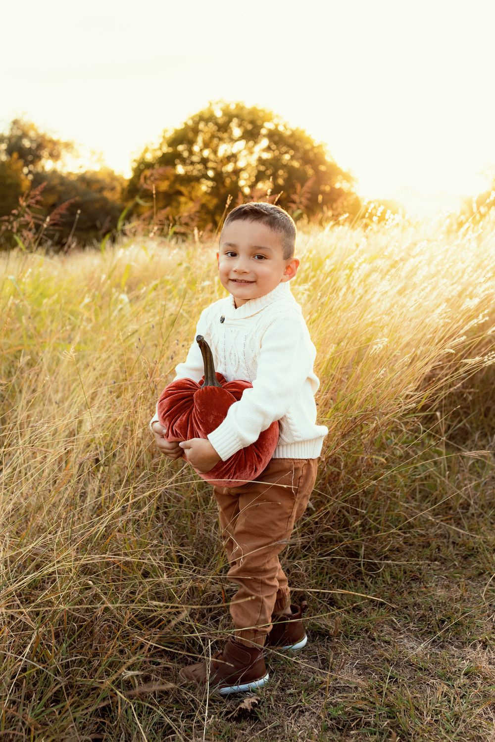 Young child in white sweater and brown pants holds football in golden sunlit field at sunset.