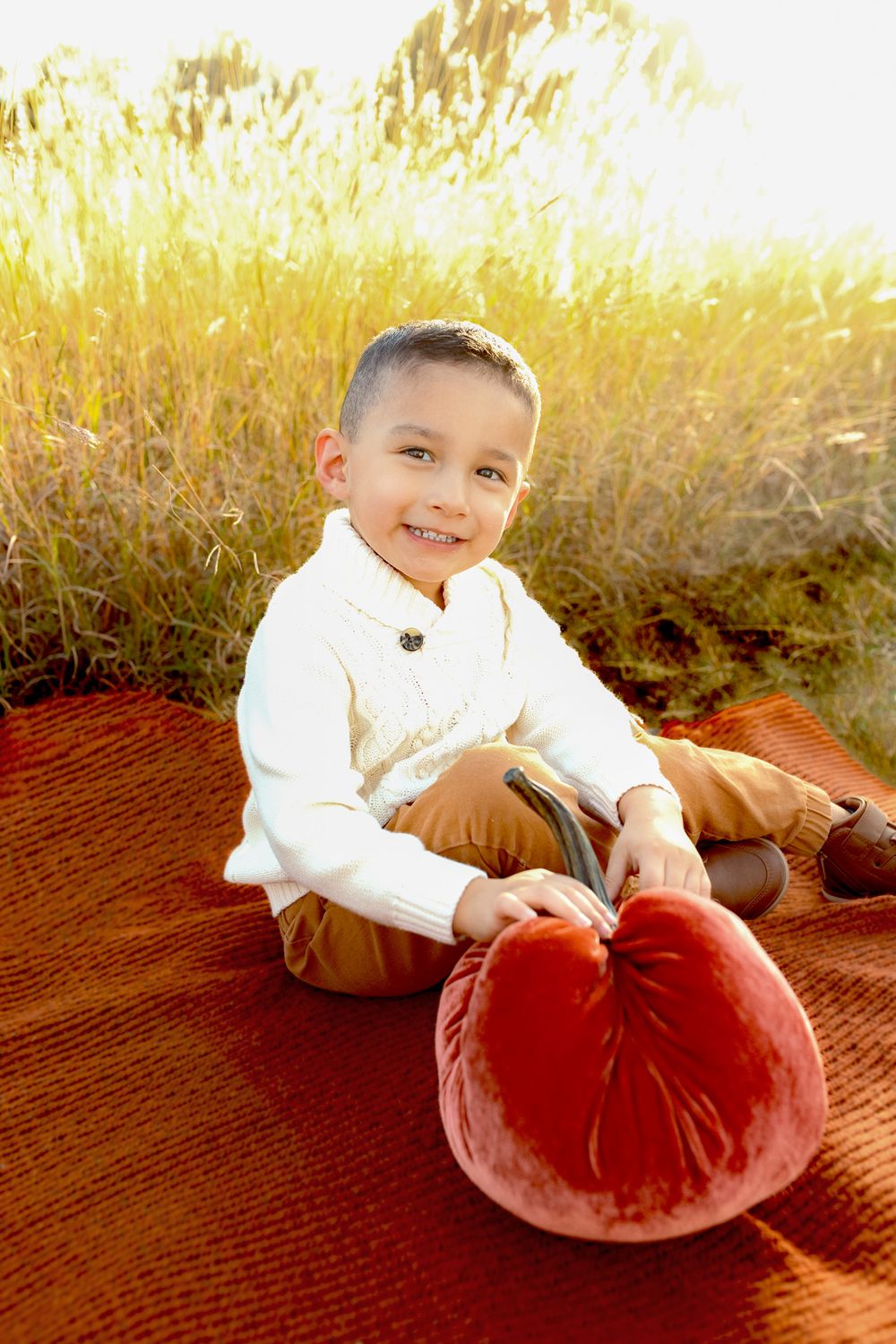 Child in white sweater sits on orange blanket with large red velvet pumpkin at sunset.