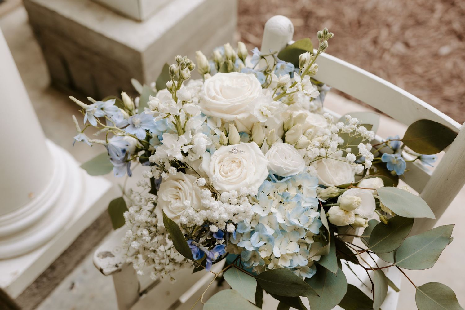 Elegant wedding bouquet featuring white roses, blue hydrangeas, and baby's breath arranged on a white wooden surface.