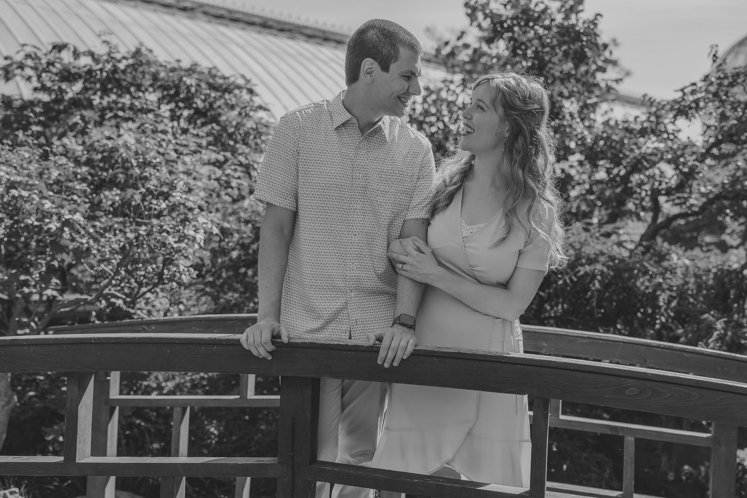 A couple stands together on a curved bridge in a romantic black and white outdoor setting.
