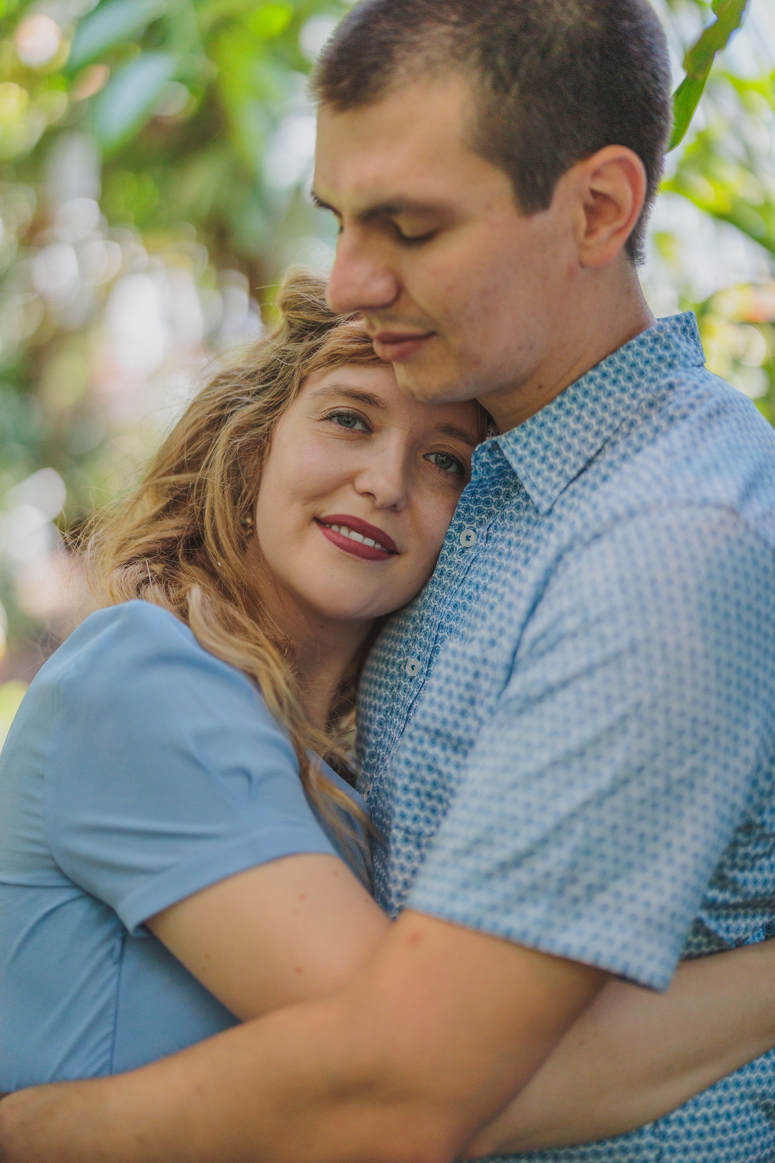 A couple embraces tenderly outdoors in blue clothing against a soft bokeh background of greenery.
