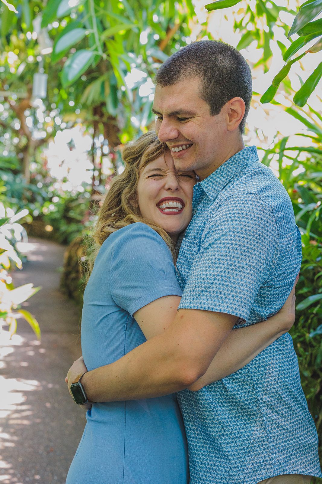 A joyful couple embraces and laughs together while standing in a garden path surrounded by green foliage.