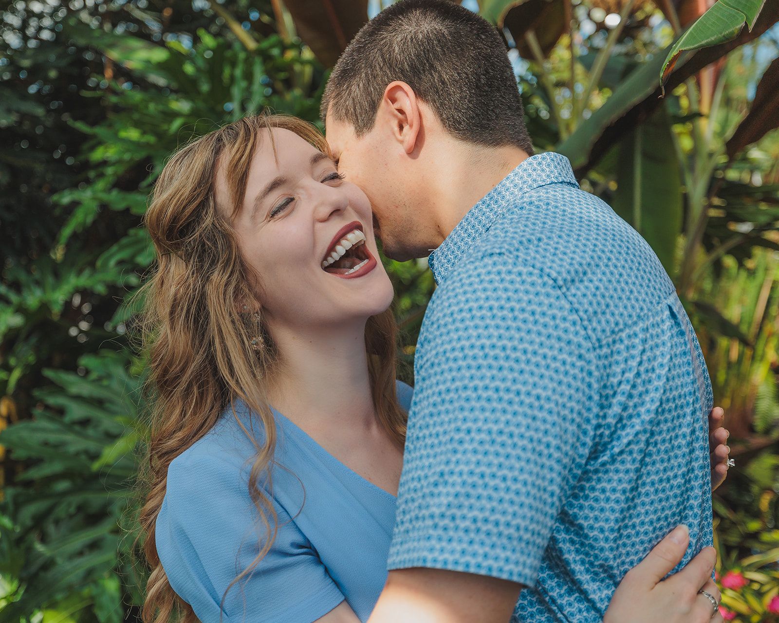 A joyful couple in matching blue outfits embrace and laugh together in front of lush green foliage.