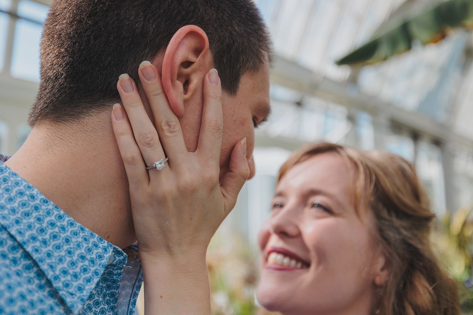 A couple shares an intimate moment outdoors, with one person covering their face while the other smiles warmly.