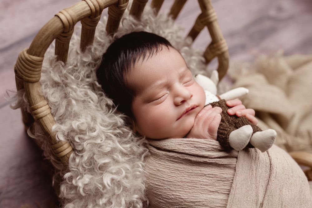 A peaceful sleeping newborn wrapped in beige fabric rests in a wooden basket on fluffy white fur.