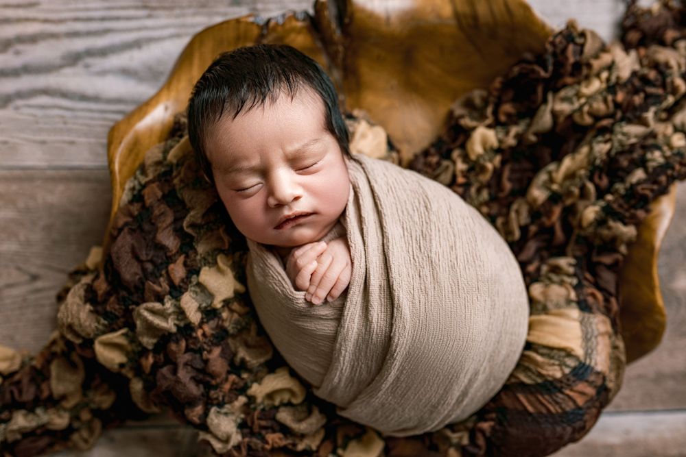 Peaceful sleeping newborn baby wrapped in beige swaddle blanket on brown textured backdrop during professional photoshoot.