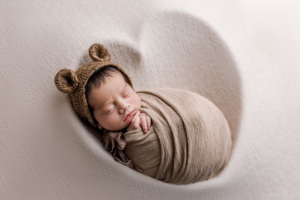 A sleeping newborn swaddled in beige fabric wearing a brown knitted bear hat rests in a heart-shaped frame.