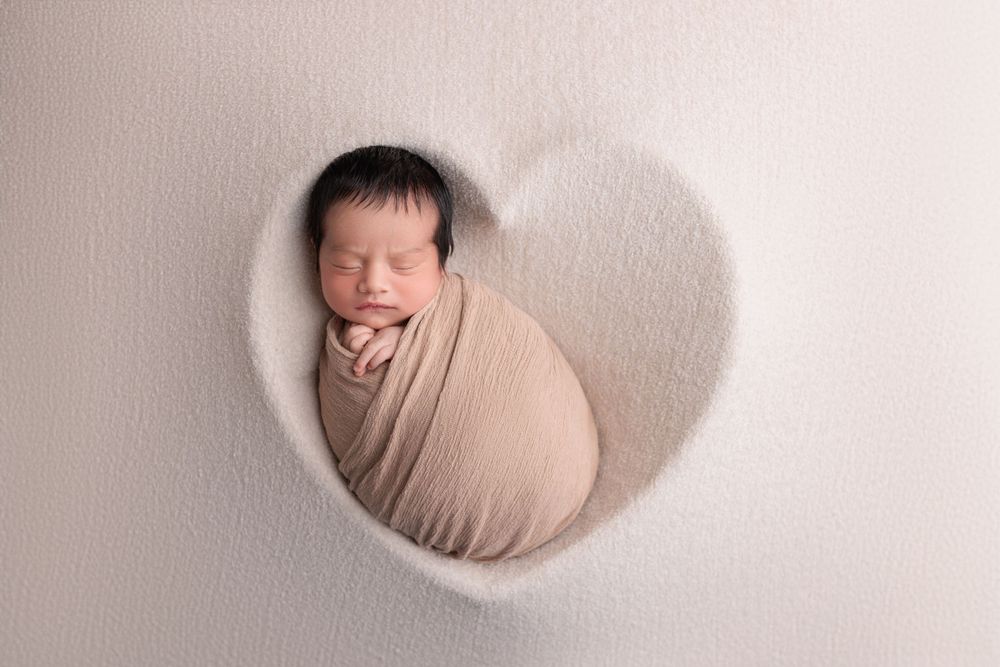 A sleeping newborn baby swaddled in beige wrap positioned in a white heart-shaped frame during photography session.