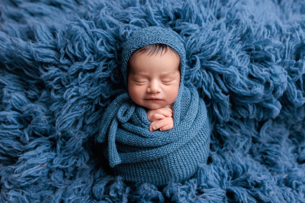 Sleeping newborn wrapped in blue knit swaddle rests peacefully on fluffy blue blanket during professional photography session.