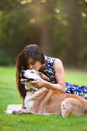 Woman in floral dress kissing her happy golden dog on grass in a sunny park.