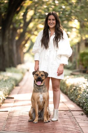 Cheerful mixed-breed dog sits on brick pathway next to someone wearing white dress and silver boots in sunny garden setting.
