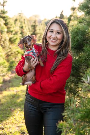 Young woman in red sweater holding small dachshund puppy wearing plaid outfit among evergreen trees in outdoor setting.