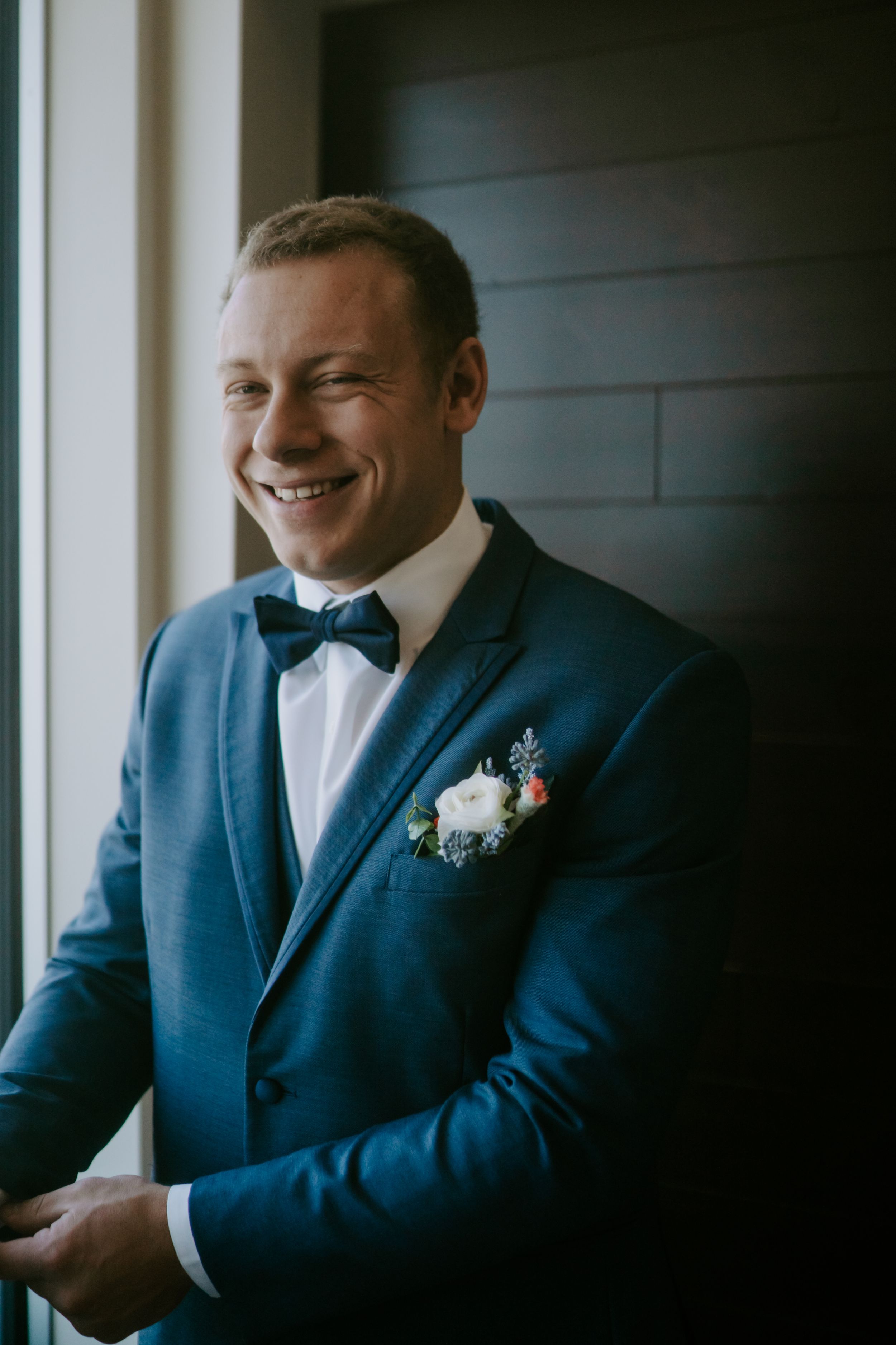 Groom in navy blue suit with boutonniere smiles by window in natural light.