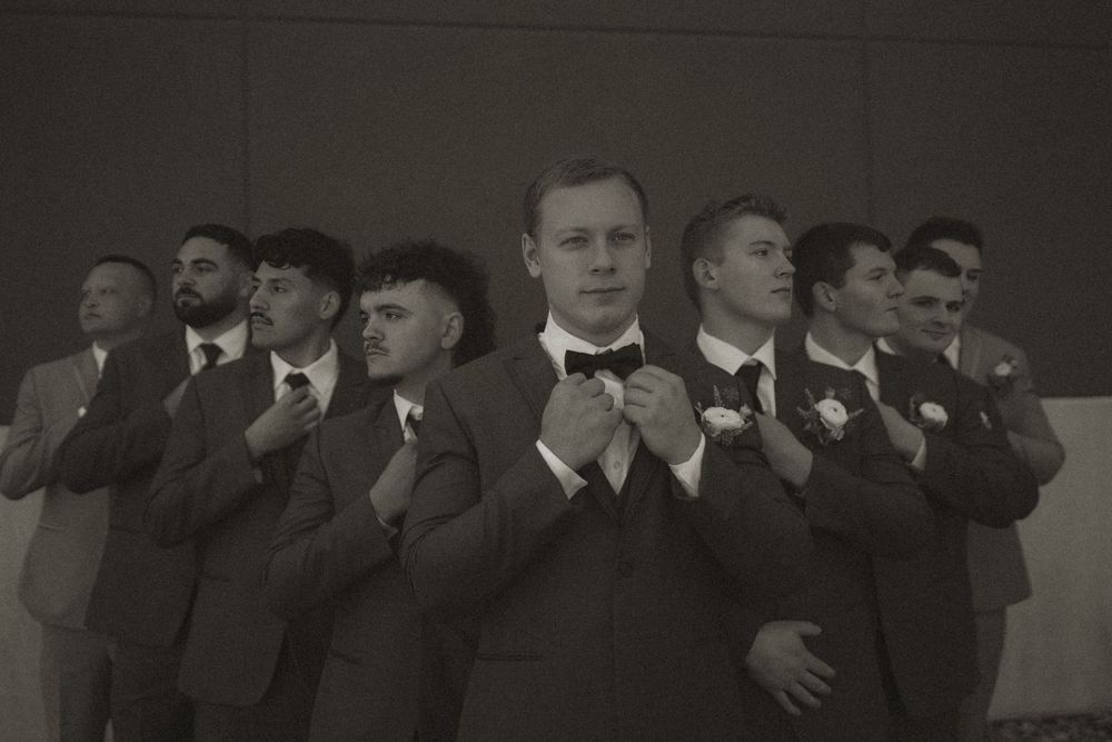 Wedding party in dark suits lined up against grey wall for formal group photo.