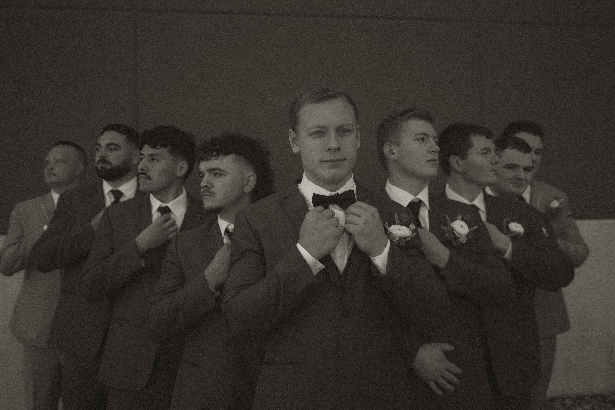 Wedding party in dark suits lined up against grey wall for formal group photo.