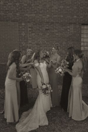 Wedding party in neutral dresses poses against brick wall outdoors.