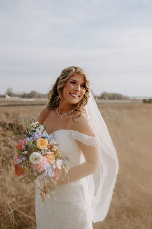 A bride in a white gown holds a colorful bouquet of flowers against a rustic outdoor backdrop.