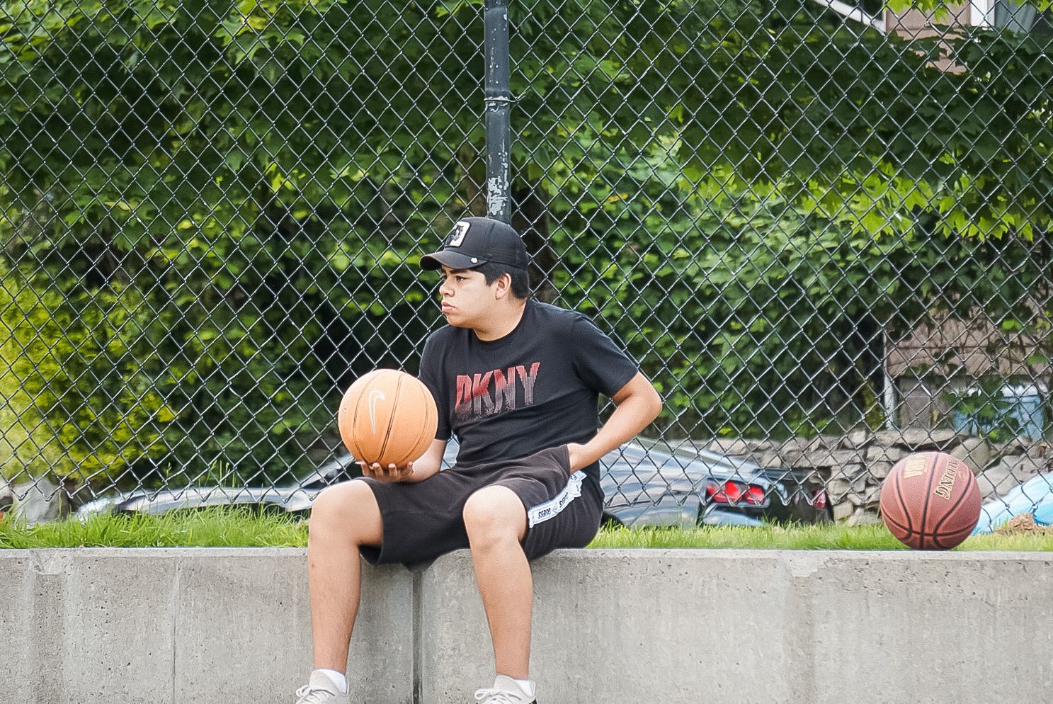 A person in a black t-shirt sits on a concrete ledge at a basketball court while holding an orange basketball.