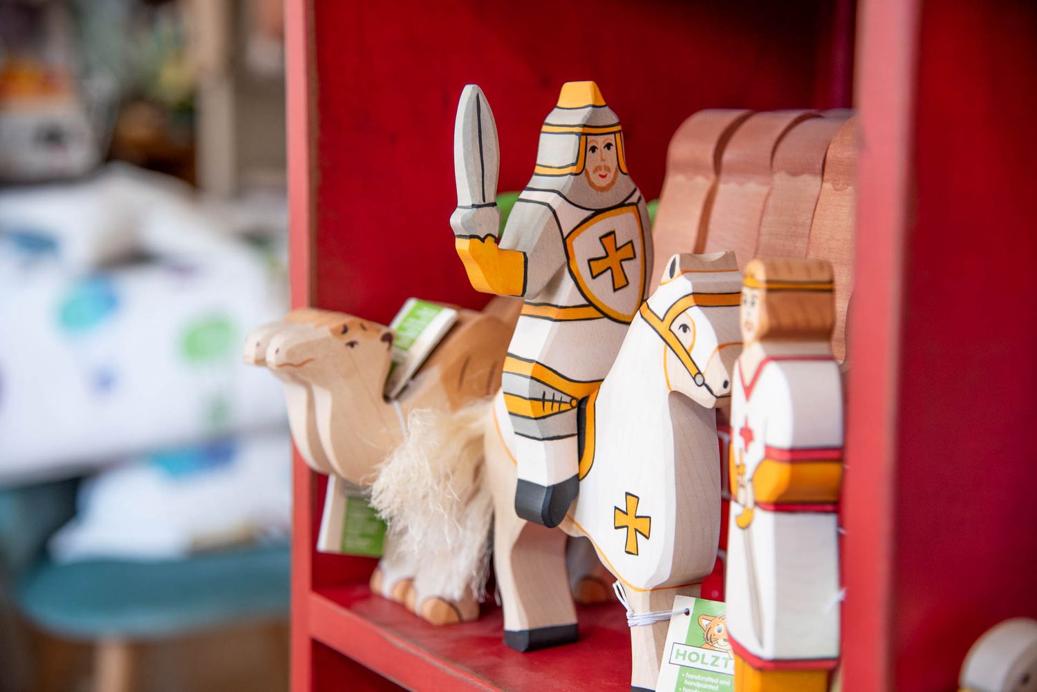 Small wooden knight toy with sword and shield mounted on white horse displayed on red shelf background.
