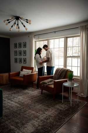 A romantic couple embraces in a sunlit living room with brown carpet, mid-century modern furniture and retro light fixture.