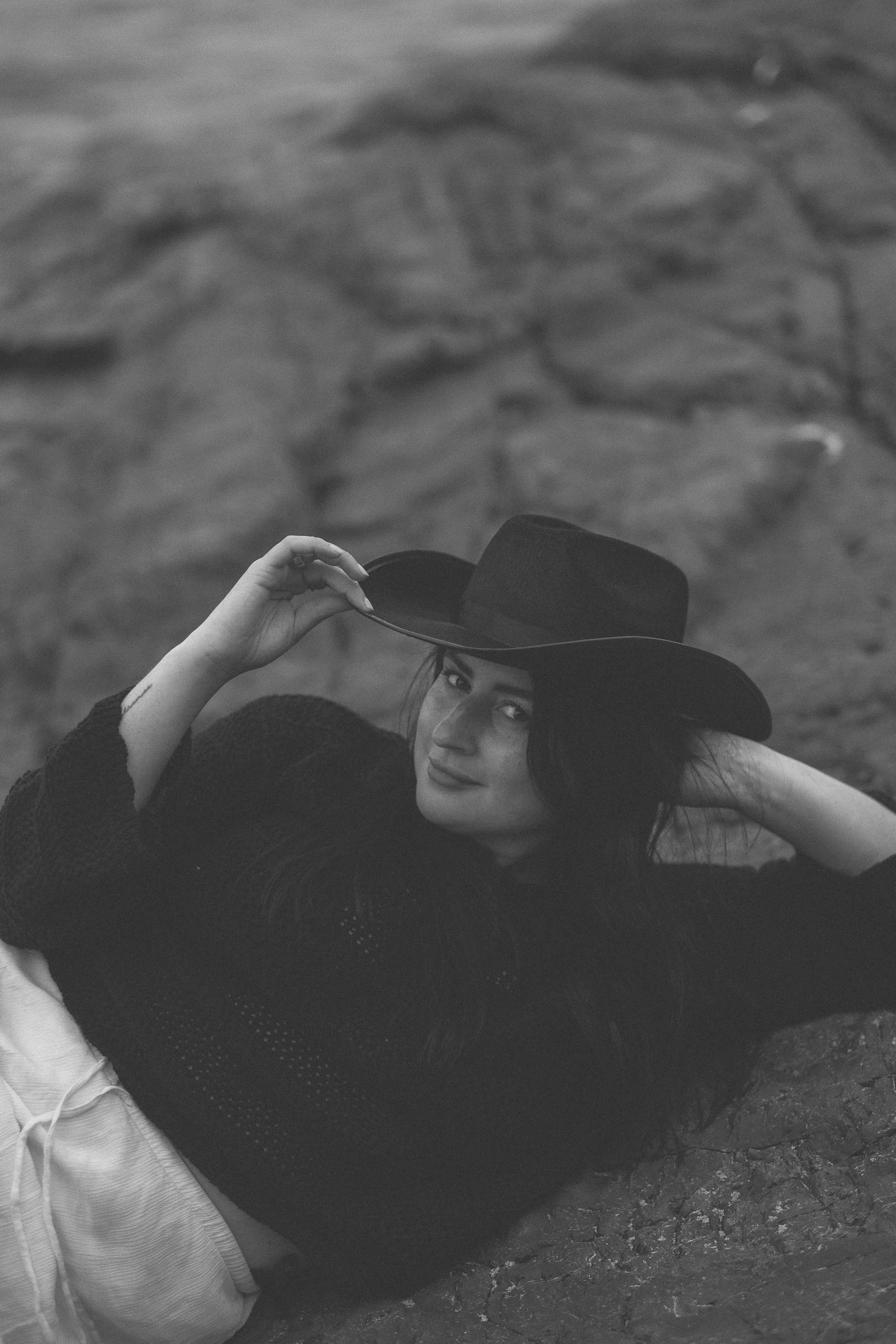 A black and white portrait of someone reclining on the ground wearing a dark cowboy hat and sweater.