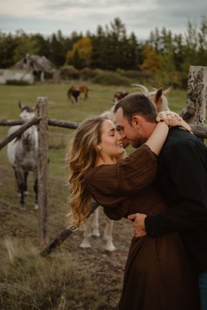 A romantic couple embraces in a rustic field with horses grazing in the background during golden hour.