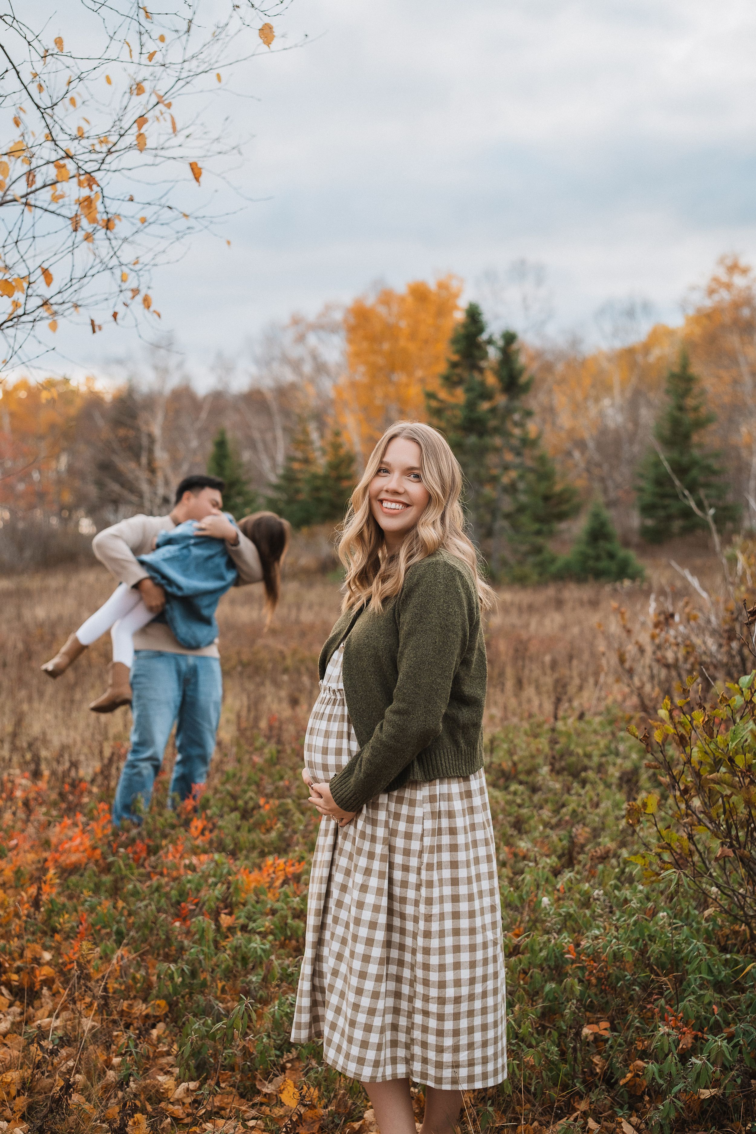 A person in a gingham dress and olive cardigan stands in an autumn field with orange foliage and bare trees.