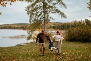 Family enjoying an autumn day by the lake, swinging child between them on grass covered with fallen leaves.