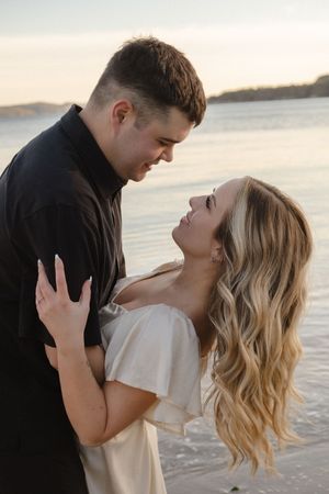 A romantic couple embraces on a beach at sunset, with waves and mountains in the background.