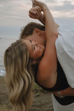 A romantic couple embraces and shares an intimate kiss against a cloudy sky backdrop.