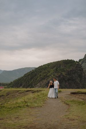 Two figures stand on a scenic mountain overlook with dramatic cloudy skies and green hills stretching into the distance.