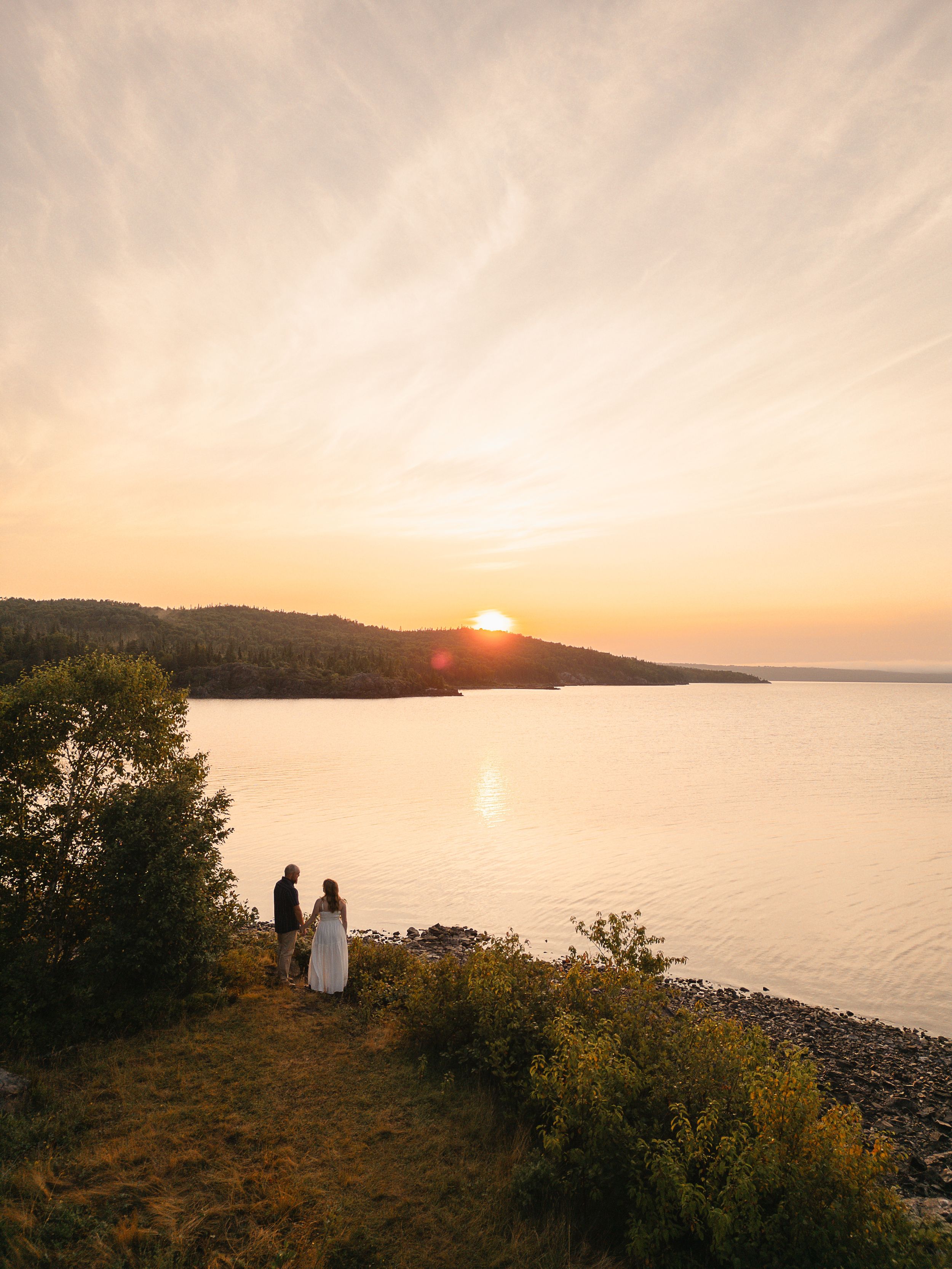 Two silhouettes stand on a rocky shoreline at sunset as golden light reflects off a tranquil lake surrounded by hills.