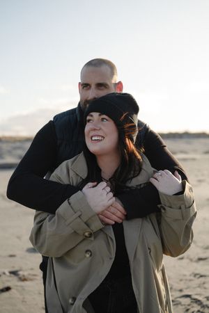 A couple embraces on a beach during golden hour, with the person in front wearing a beige jacket and black beanie.