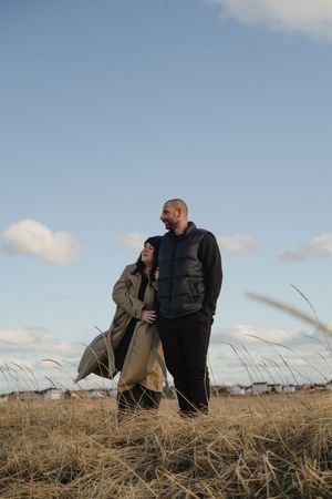 A couple stands together in a field of dry grass against a blue sky with white clouds on a sunny day.