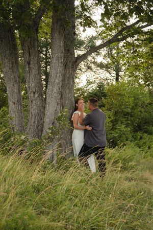 A couple shares a romantic moment together in tall grass beneath large trees at sunset.