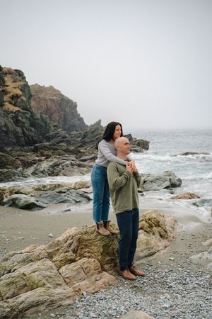 Couple embracing on rocky coastal beach with ocean waves and misty cliffs in the background.