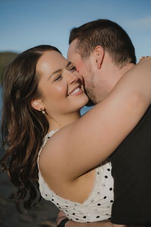 A smiling woman in a white polka dot dress embraces a man kissing her cheek outdoors under a blue sky.