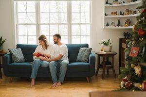 Couple sitting together on blue couch in bright living room with large windows and Christmas tree decorations.