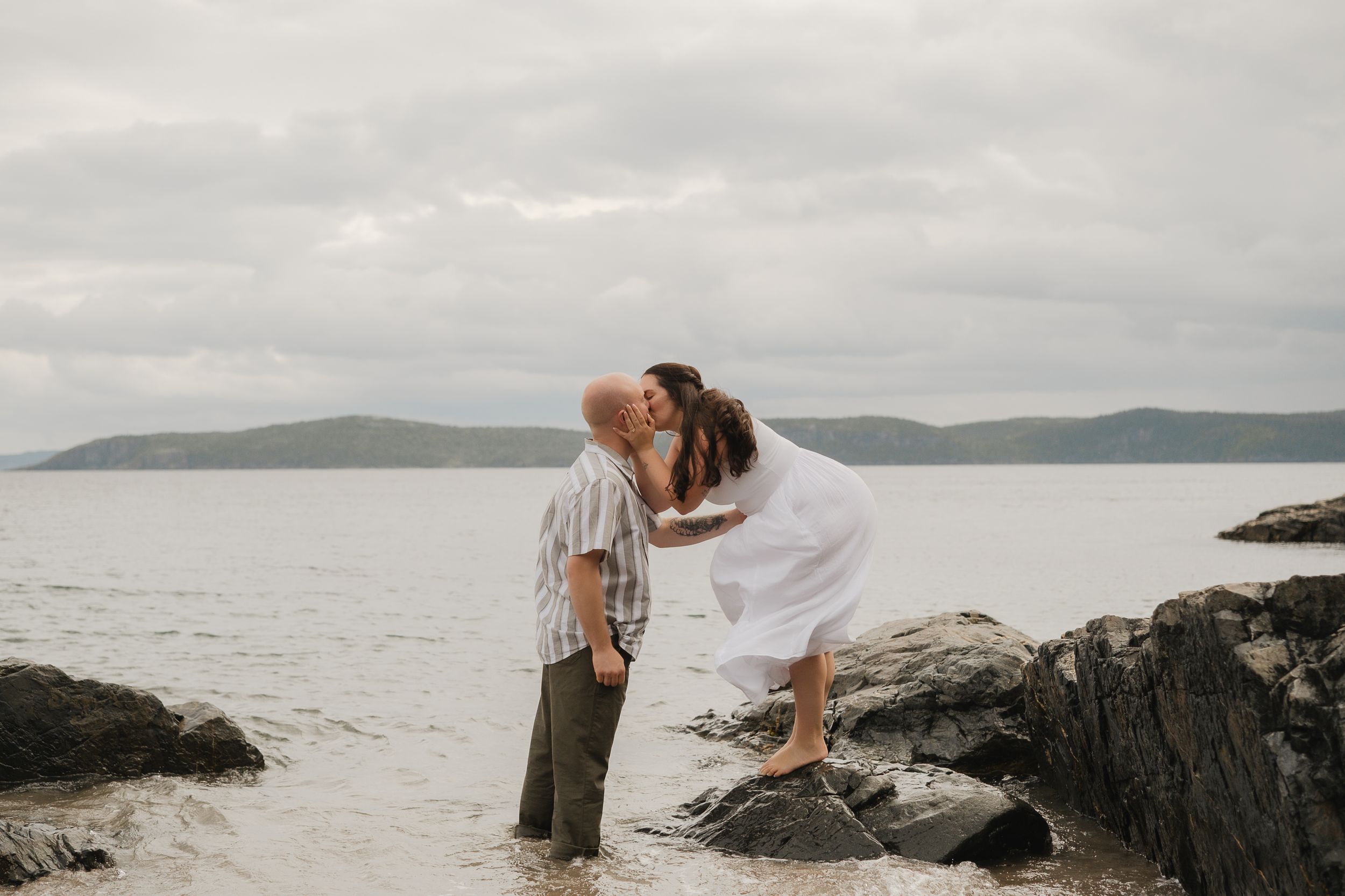 A couple shares a romantic moment on rocky coastal cliffs overlooking a serene ocean view on an overcast day.
