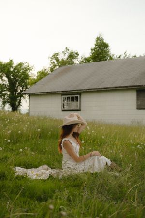 A person in a white dress and sun hat sits peacefully in a lush summer meadow beside a white house.