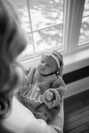 A newborn baby sleeping peacefully near a window in a cozy gray sweater, captured in black and white photography.