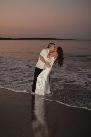 A romantic couple in white attire share a passionate embrace on a beach at sunset with ocean waves in the background.