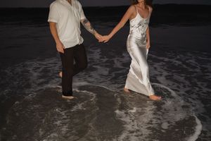 A couple in wedding attire walks together in shallow ocean waves at night during a beach photo session.