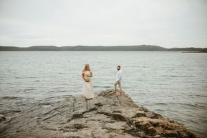 Couple in white attire standing on rocky shoreline with calm lake and distant hills backdrop.