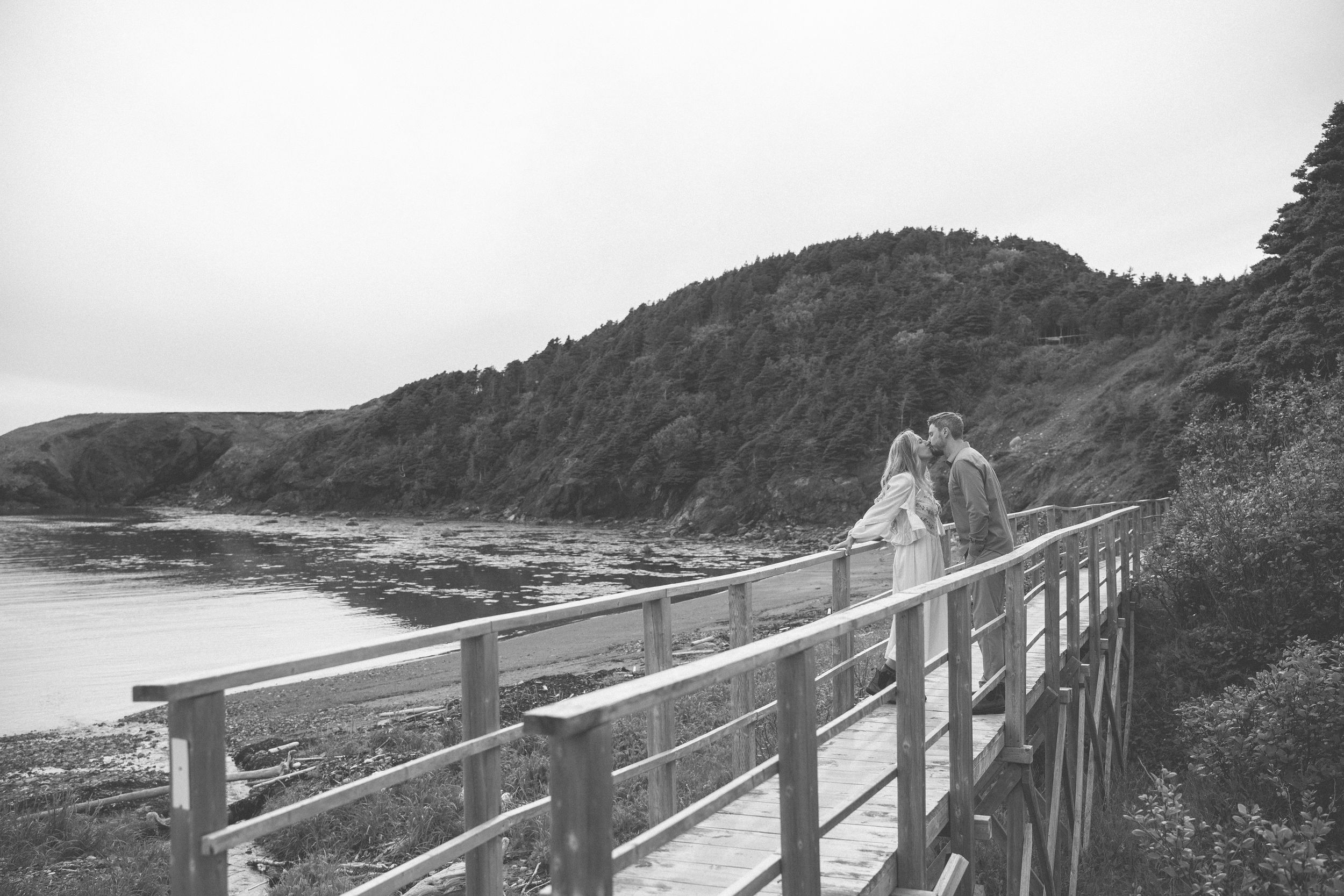 Black and white photo of a couple on a wooden boardwalk by a scenic coastal bay with lush green hills.