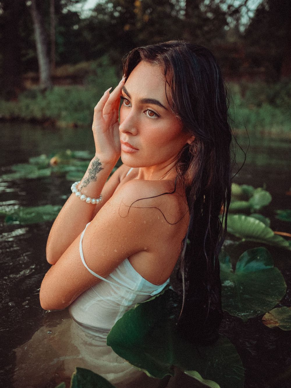 A woman in a white top poses thoughtfully by a pond with lily pads in a romantic outdoor photoshoot.