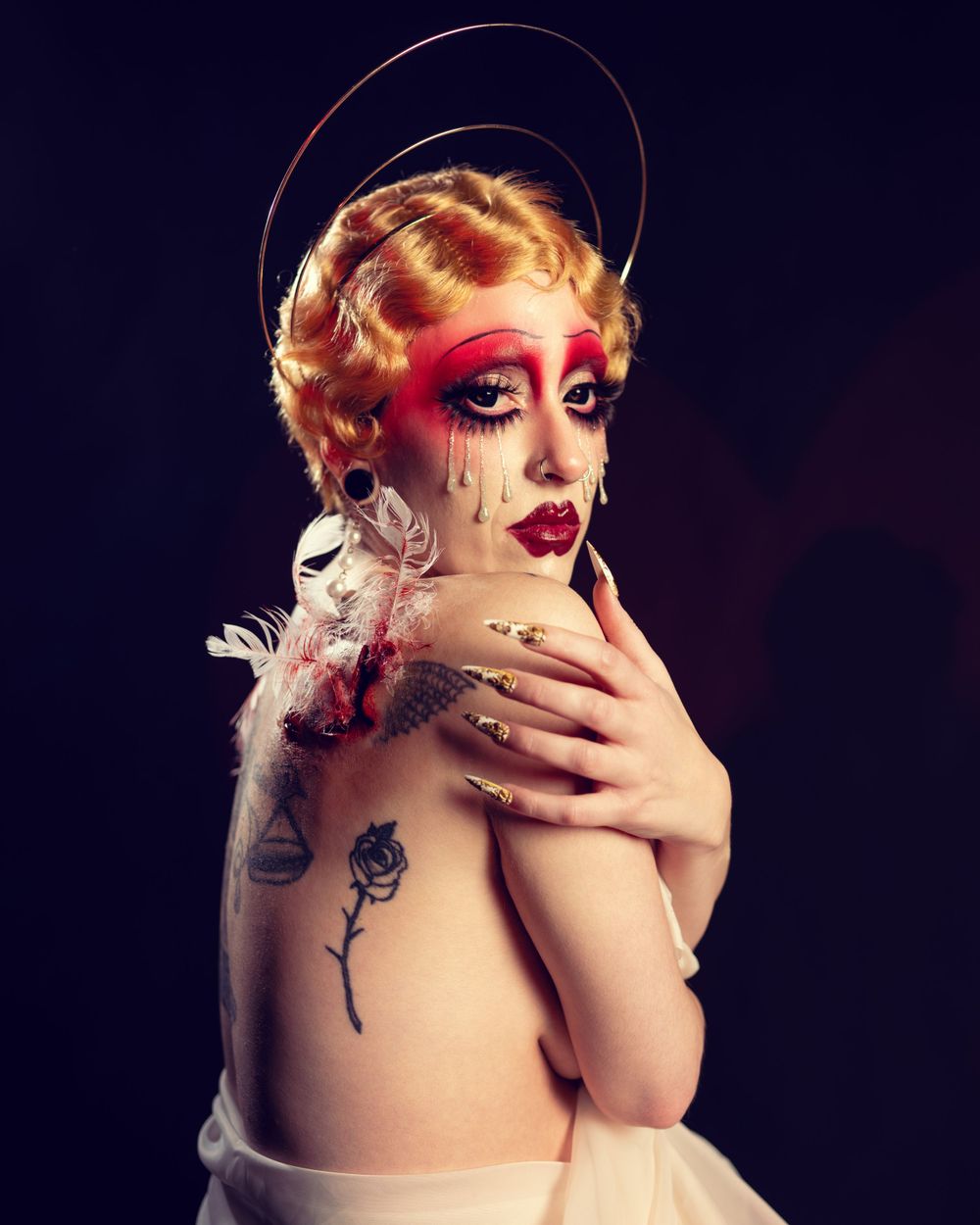 A dramatic artistic portrait in dark tones featuring dramatic makeup, finger-waved blonde hair, and rose tattoo on bare shoulder.