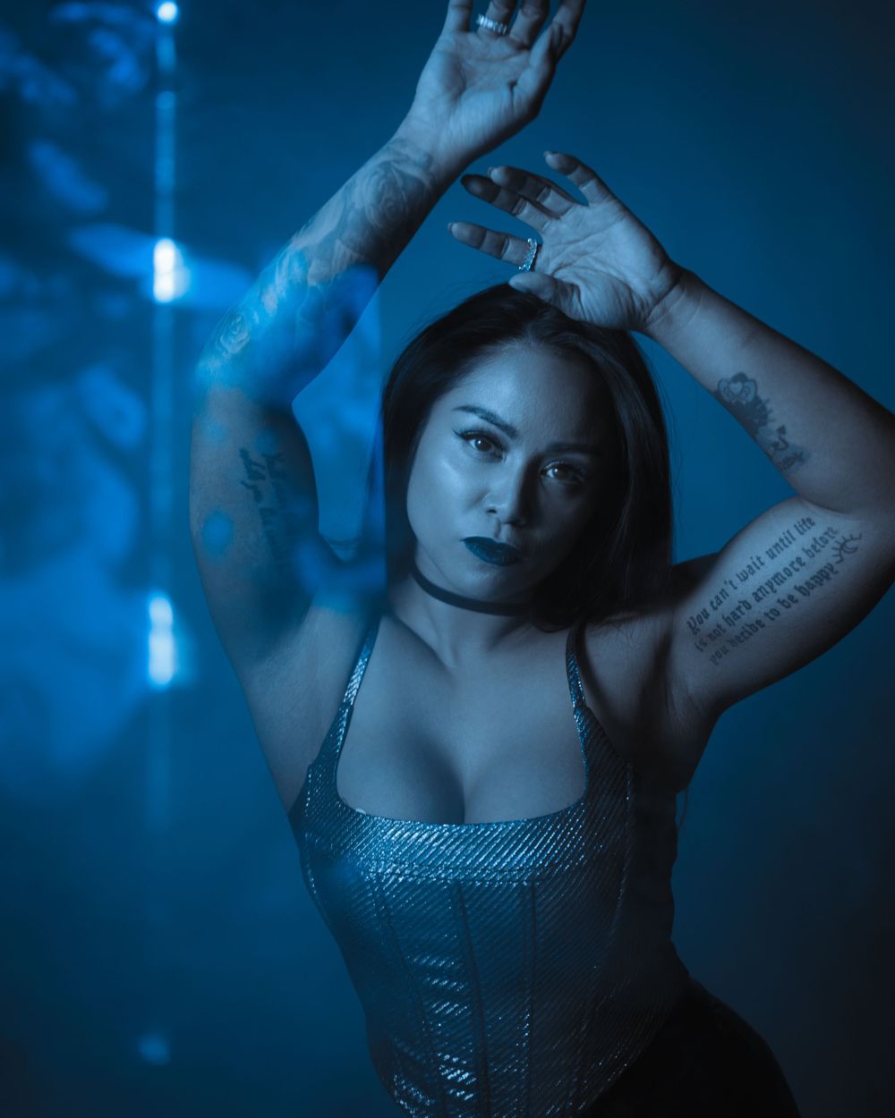 Moody portrait with dramatic blue lighting shows a person in sparkly top posing with raised arms against dark studio photography background.